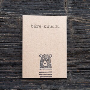 May include: A brown card with the text "bäre-knuddu" printed on it. A black and white illustration of a bear's face is printed at the bottom of the card.