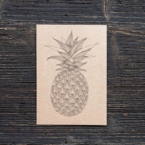 May include: A pen and ink drawing of a pineapple on a kraft paper card. The pineapple illustration is centered against a dark wood grain background. The drawing shows the fruit's spiky leaves and textured skin. The card is rectangular.