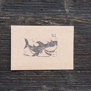 May include: A brown card with a black and white illustration of a shark with the word "hi" printed above it.