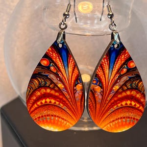 May include: A pair of teardrop-shaped earrings with a vibrant, abstract design. The earrings feature a combination of orange, red, and blue hues, with intricate patterns. The earrings are suspended from silver-colored ear wires, showcasing a unique and artistic style.