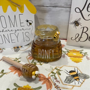 May include: A glass honey jar with a yellow and black bee accent, filled with honey. A wooden honey dipper and a decorative sign with the words "Home where your honey is" are also visible. The jar has the word "HONEY" printed on it.