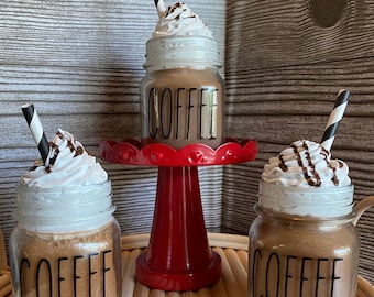 May include: Three decorative coffee jars with whipped cream and chocolate drizzle. Each jar has the word "COFFEE" in black lettering. One jar sits on a red pedestal stand, and all have black and white striped straws.