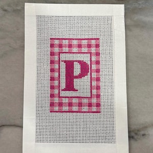 May include: A needlepoint canvas featuring a white background with a pink gingham pattern and a framed pink letter "P". The canvas is rectangular with a white border, perfect for a personalized craft project.