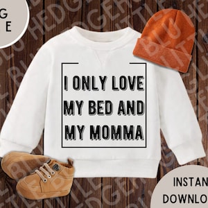 以下が含まれることがあります： 白いスウェットシャツに、黒い文字で「ベッドとママしか愛してない」と書かれています。スウェットシャツは木製の背景に置かれ、茶色のビーニー帽と茶色の靴が置かれています。