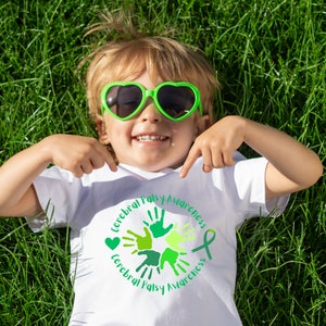 Puede incluir: Un ni&ntilde;o peque&ntilde;o con una camiseta blanca con un dise&ntilde;o circular verde que dice "Cerebral Palsy Awareness" y huellas de manos verdes. El ni&ntilde;o lleva gafas de sol verdes en forma de coraz&oacute;n y est&aacute; tumbado de espaldas en un campo de hierba.