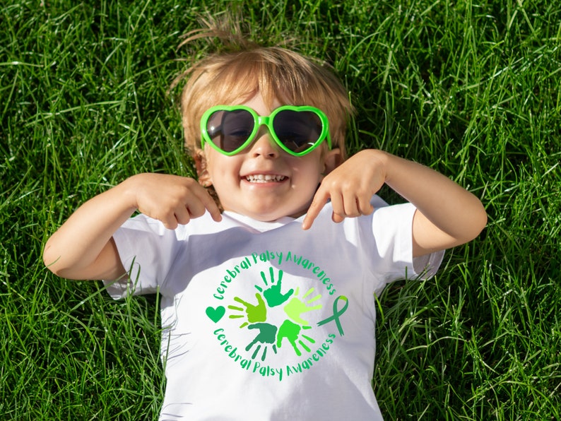 Puede incluir: Un ni&ntilde;o peque&ntilde;o con una camiseta blanca con un dise&ntilde;o circular verde que dice "Cerebral Palsy Awareness" y huellas de manos verdes. El ni&ntilde;o lleva gafas de sol verdes en forma de coraz&oacute;n y est&aacute; tumbado de espaldas en un campo de hierba.