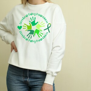Puede incluir: Sudadera blanca con una cinta verde y huellas de manos en un c&iacute;rculo. El texto "Cerebral Palsy Awareness" est&aacute; escrito alrededor del c&iacute;rculo.