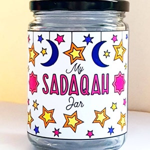May include: A glass jar with a black lid and a white label that says "My Sadaqah Jar". The label is decorated with stars and crescent moons in various colors.