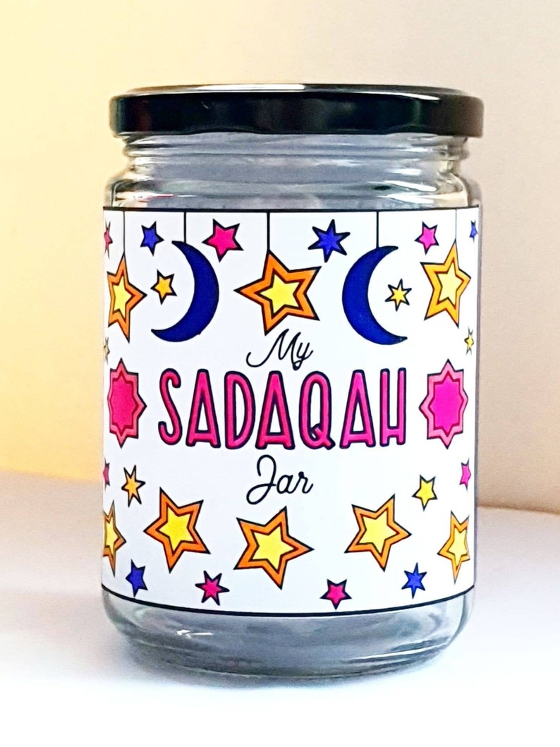 May include: A glass jar with a black lid and a white label that says "My Sadaqah Jar". The label is decorated with stars and crescent moons in various colors.