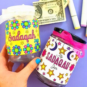 May include: Two glass jars with colorful labels that say "My Sadaqah Jar". One jar has a yellow background with blue, pink, and green stars and geometric shapes. The other jar has a white background with purple, pink, and yellow stars and crescent moons. Both jars have pink ribbons tied around the lids.
