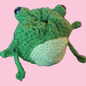 May include: Green crocheted frog toy with a light green belly. The frog has black eyes and a textured surface.
