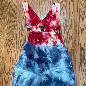 May include: Tie-dye denim overalls with adjustable straps and a red, white, and blue color scheme. The overalls have a  "Polo Jeans Co. Ralph Lauren" label on the front.