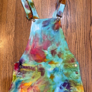 May include: Tie-dye denim overalls with a distressed hem and a light blue pocket. The overalls are a mix of blue, green, purple, orange, and yellow.