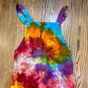 May include: Tie-dye denim overalls with a rainbow swirl pattern in shades of red, orange, yellow, green, blue, and purple. The overalls have adjustable straps and a frayed hem.
