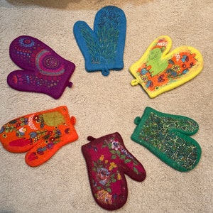 May include: Six colorful oven mitts with different patterns. The mitts are made of felt and have a loop for hanging. The patterns include flowers, a frog, and geometric designs.
