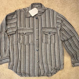 May include: A long-sleeved button-down shirt with vertical stripes in shades of gray, brown, and white. The shirt has two chest pockets and a collar. The buttons are a dark color. A small tag is attached to the collar.