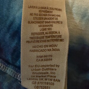 Puede incluir: Primer plano de una etiqueta de tela con instrucciones de cuidado en franc&eacute;s y espa&ntilde;ol. La etiqueta incluye el texto "HECHO EN INDIA" y "FABRICADO NA INDIA". La etiqueta est&aacute; cosida a una tela te&ntilde;ida anudada azul y blanca.