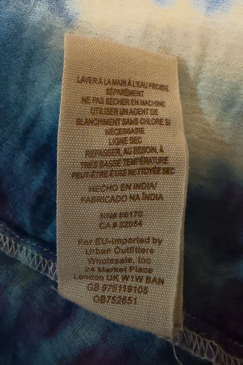 Puede incluir: Primer plano de una etiqueta de tela con instrucciones de cuidado en franc&eacute;s y espa&ntilde;ol. La etiqueta incluye el texto "HECHO EN INDIA" y "FABRICADO NA INDIA". La etiqueta est&aacute; cosida a una tela te&ntilde;ida anudada azul y blanca.