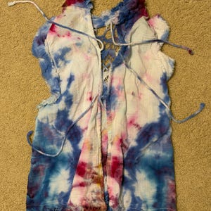 Puede incluir: Una blusa te&ntilde;ida con tintes de azul, rosa y blanco. La prenda presenta un frente con cordones y bordes deshilachados. La blusa est&aacute; hecha de una tela ligera.