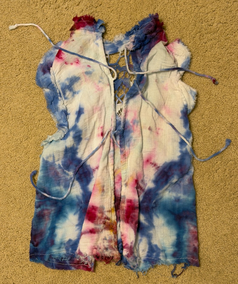 Puede incluir: Una blusa te&ntilde;ida con tintes de azul, rosa y blanco. La prenda presenta un frente con cordones y bordes deshilachados. La blusa est&aacute; hecha de una tela ligera.