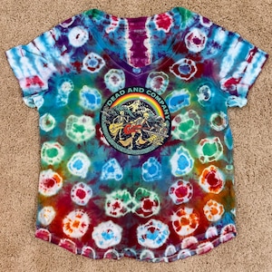 May include: A tie-dye V-neck t-shirt with a vibrant pattern of blues, greens, reds, and purples. The shirt features a circular patch with the text "DEAD AND COMPANY" and a graphic of skeletons playing instruments against a mountain backdrop.