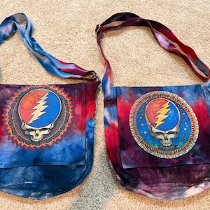 May include: Two tie-dye shoulder bags. Each bag features a Steal Your Face skull design in a circle with a red and orange top, blue lightning bolt, and a skull. The bags have blue, red, and purple tie-dye patterns and matching straps.