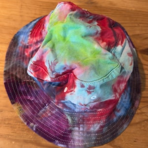 May include: A tie-dye bucket hat with a colorful pattern of red, blue, green, and purple. The hat has a wide brim and a flat crown.