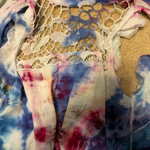 Puede incluir: Una blusa te&ntilde;ida con un panel trasero de crochet blanco. La blusa presenta una mezcla de colores azul, rosa y blanco. La tela tiene un dise&ntilde;o de borde deshilachado y deshilachado. La blusa est&aacute; hecha de un material ligero.