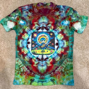 May include: A colorful tie-dye t-shirt with a vibrant design. The shirt features a central graphic of a skeleton playing a guitar under a sun and moon. The shirt's tie-dye pattern includes shades of green, blue, red, and purple.