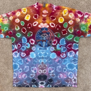 May include: A tie-dye t-shirt with a circular graphic featuring a skeleton wearing an Uncle Sam hat and bow tie. The shirt has a colorful pattern of red, yellow, blue, and green spots.