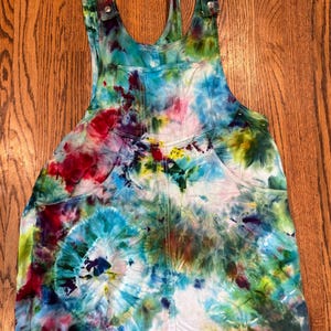 May include: Tie-dye denim overalls with adjustable straps and pockets. The overalls are a light blue color with swirls of red, green, yellow, and purple.