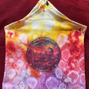 May include: A tie-dye halter top with a round graphic in the center. The top features a vibrant blend of yellow, orange, pink, and purple hues. The graphic reads "Dead and Company In Concert" and depicts a colorful landscape.