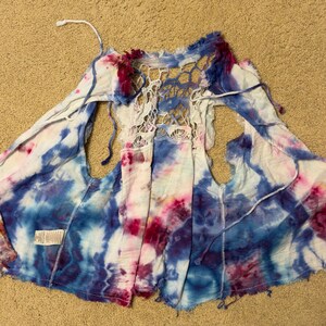 Puede incluir: Un chaleco te&ntilde;ido anudado en tonos de azul, blanco y rosa. La parte trasera presenta un dise&ntilde;o de ganchillo blanco de tejido abierto. El chaleco tiene un frente fluido y abierto y est&aacute; hecho de una tela ligera. Los bordes est&aacute;n deshilachados.