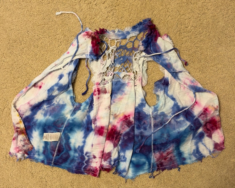 Puede incluir: Un chaleco te&ntilde;ido anudado en tonos de azul, blanco y rosa. La parte trasera presenta un dise&ntilde;o de ganchillo blanco de tejido abierto. El chaleco tiene un frente fluido y abierto y est&aacute; hecho de una tela ligera. Los bordes est&aacute;n deshilachados.