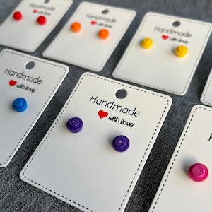May include: A collection of colorful stud earrings in various colors, including red, orange, yellow, blue, purple, and pink. Each pair is displayed on a white card with the text "Handmade with love".