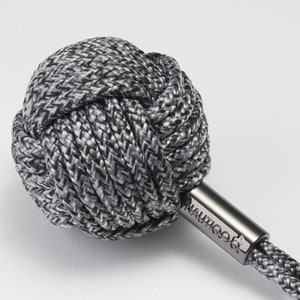 以下が含まれることがあります： グレーと白の織りボールノットが特徴の、航海をテーマにしたキーホルダー。 ノットは「Nautica」の文字が刻まれた金属バーに取り付けられ、金属製のキーリングで終わるグレーと白のロープが付いています。