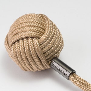 以下が含まれることがあります： ベージュ色の海事ノットキーチェーン。球形のノットは、しっかりと織られたロープで作られています。「Nauticool」という単語が入った金属チューブがノットに取り付けられています。小さなロープが銀色のキーリングに伸びています。