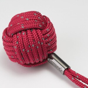 以下が含まれることがあります： 赤いグレーのロープのキーホルダーで、結び目のついたボールと銀色の金属製のリングが付いています。キーホルダーには、金属部分に「NAUTICA」という文字が刻印されています。