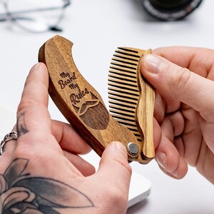 May include: A wooden folding comb with a brown wood handle and a natural wood comb. The handle has the text "My Beard My Rules" engraved on it with a small mustache icon.