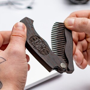 Peut inclure: Un peigne à barbe en bois brun foncé avec un design pliable. Le peigne est orné d'un motif gravé représentant un homme barbu avec des lunettes de soleil et une moustache. Le texte "Beard" est gravé au-dessus du visage de l'homme.