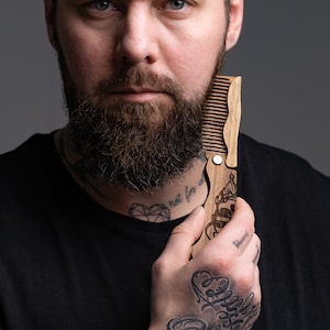 May include: A man with a beard and tattoos holds a wooden comb with a decorative design. The comb has a metal hinge and a small metal loop at the top.