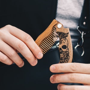 Peut inclure: Un peigne à barbe en bois avec un motif de crâne. Le peigne est en bois brun clair et a une conception articulée. Le crâne a une barbe et une moustache. Le peigne est ouvert, montrant les dents.