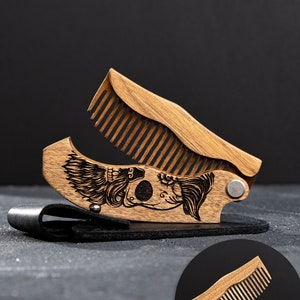 May include: A wooden folding comb with an engraved design of a mythical creature. The comb is open, showing the teeth, and sits on a black leather holder. The comb is made of light brown wood and has a metal hinge. The name "Mark" is engraved on the comb.