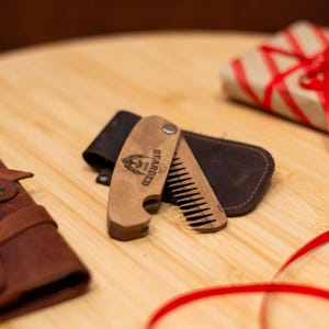 May include: A wooden beard comb and bottle opener with a leather case. The comb has a wood handle and a metal bottle opener. The case is brown leather and has a flap that closes with a snap. The comb is engraved with the words "The Bearded Man" and a logo of a bearded man.