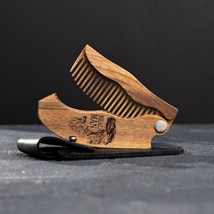 May include: A folding wooden comb in a dark brown finish, with a hinged design. The comb is engraved with the words "REAL MAN" and an illustration. It rests on a black leather case.