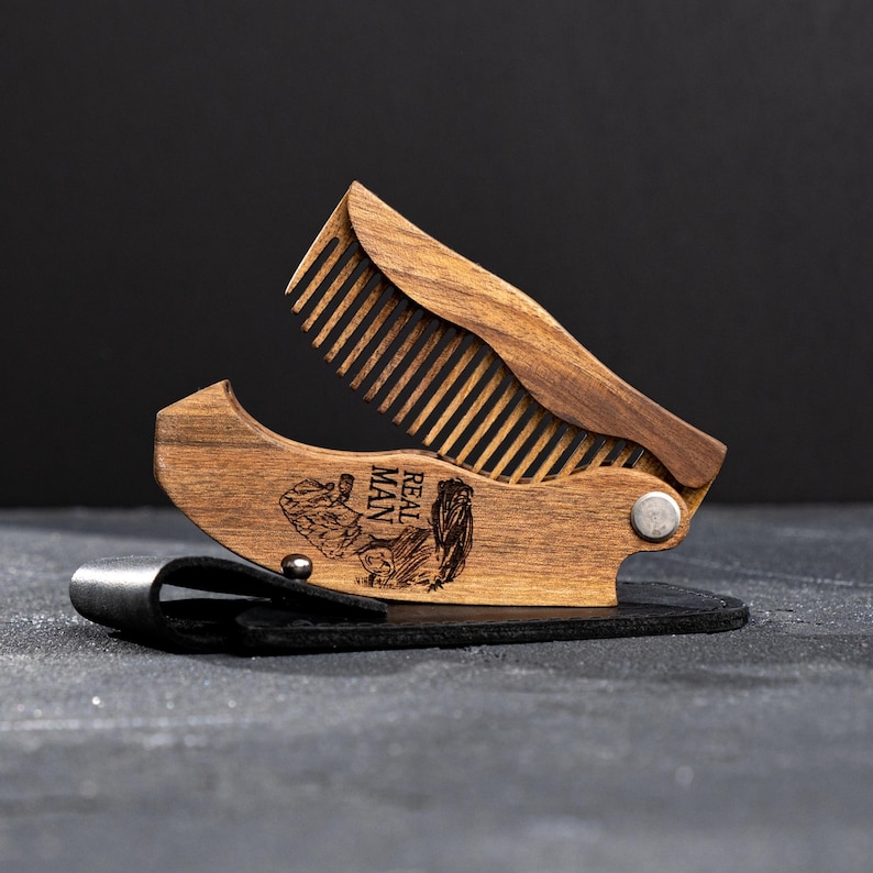 May include: A folding wooden comb in a dark brown finish, with a hinged design. The comb is engraved with the words "REAL MAN" and an illustration. It rests on a black leather case.