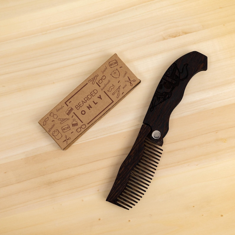May include: A dark wood folding comb with a decorative design on the handle, next to a small cardboard box with the text "BEARDED ONLY" and various beard-related illustrations. The comb is open, showing the teeth.