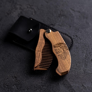 May include: A wooden folding beard comb with the words "REAL MAN" and a man's face graphic engraved on the handle. The comb is open, revealing the teeth, and rests on a black leather case.