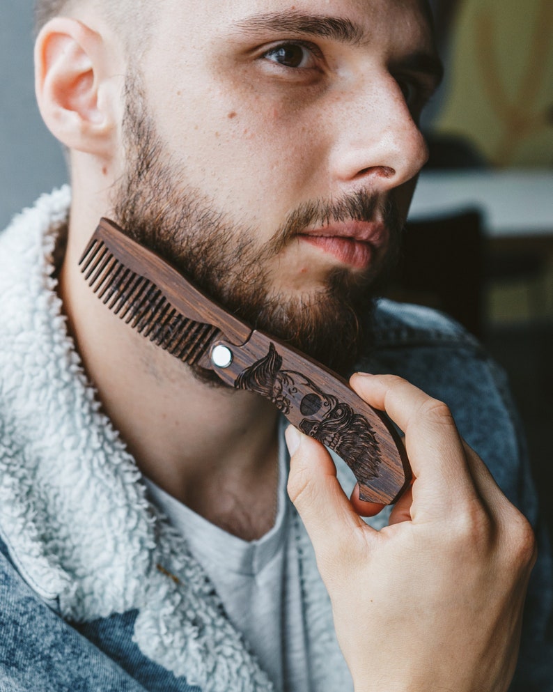 May include: A dark brown wooden beard comb featuring an engraved skull with wings. The comb is held in a hand, highlighting the detailed design and the natural grain of the wood. The comb is approximately 15 cm in length.