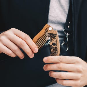 May include: A wooden folding beard comb with a skull design. The comb is made of light brown wood and features a detailed engraving of a skull with a beard and hair. The comb is held open, revealing the teeth of the comb.